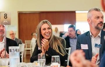 Business event attendees seated at round tables listening to a speaker, with one person in the foreground clapping and glasses and table settings visible
